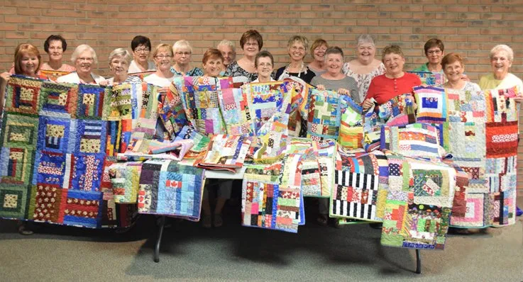 A group of members in front of a brick wall holding up multiple quilts that are covering other quilts on a table
